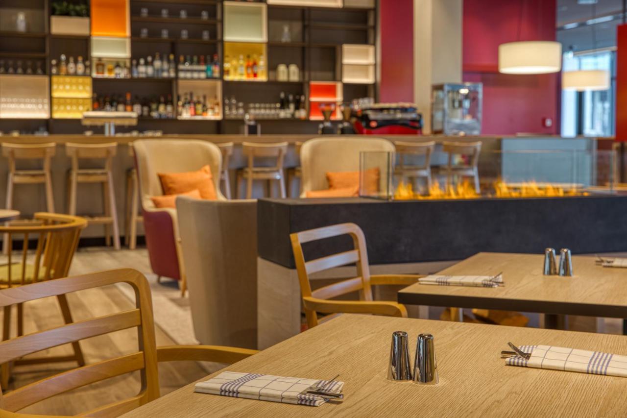 Intercityhotel Berlin Airport Ber Terminal 1 & 2 Schönefeld Exterior foto The photo shows a modern restaurant or bar interior. In the foreground, there are wooden tables set with napkins and condiments. The seating includes wooden chairs and plush seating in various colors in the background. A stylish bar area is visible w